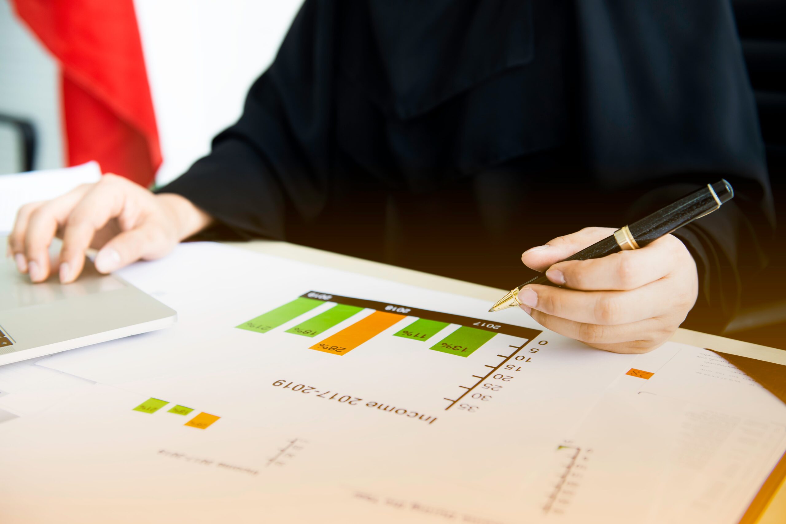 Businessperson analyzing an income graph with a pen and working on a laptop.